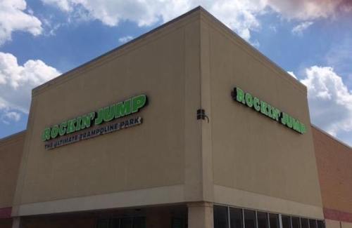 Exterior of a trampoline park building with a green sign that reads "Rockin' Jump." Bright blue sky in the background.