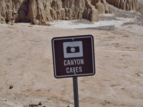A brown sign reading "Canyon Caves" with a camera icon, set against a rocky, desert landscape.