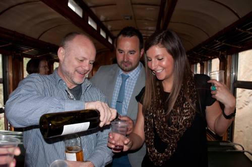 A group of three people inside a train, one pouring a drink while the others watch and hold cups.