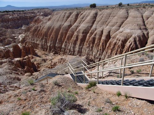 A winding staircase descends into a colorful, rugged canyon with layered rock formations under a clear blue sky.