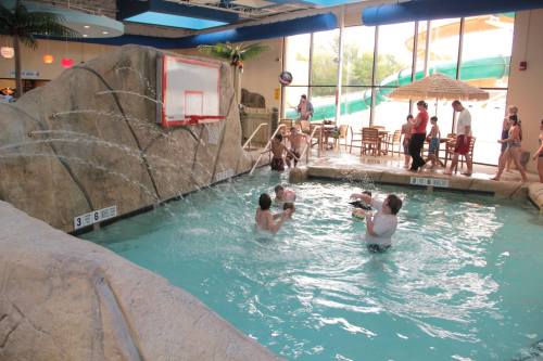 A lively indoor pool scene with people playing water basketball near a rock formation and water slides in the background.