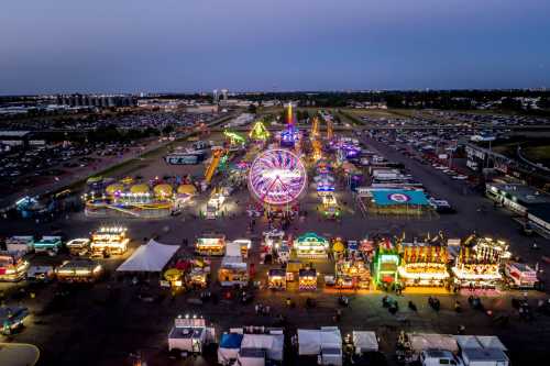 Aerial view of a vibrant carnival at dusk, featuring rides, food stalls, and colorful lights in a large parking area.