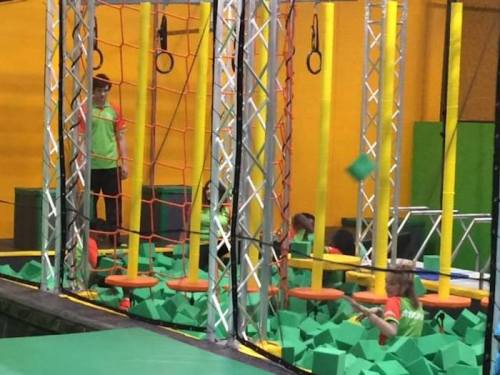 Children playing in a trampoline park, navigating obstacles and jumping into a pit filled with green foam blocks.