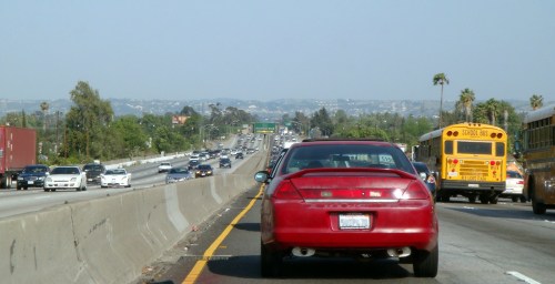 A red car on a busy highway with traffic, surrounded by greenery and a yellow school bus in the background.