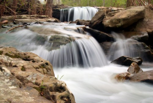 A serene waterfall cascades over rocky ledges, surrounded by lush greenery and smooth stones.