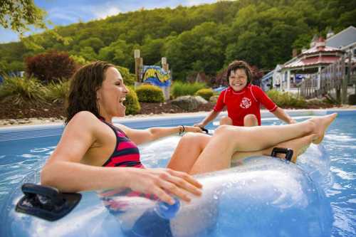 A woman and a child enjoy a fun moment on a clear inflatable tube in a water park pool, surrounded by greenery.