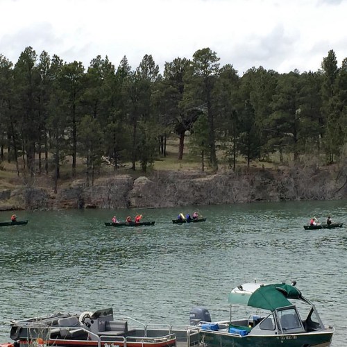 A serene lake scene with several canoes paddling, surrounded by lush green trees and rocky shores.