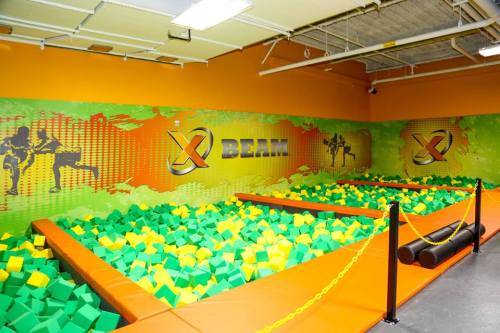 A colorful indoor trampoline park area filled with green and yellow foam blocks, featuring a vibrant wall mural.