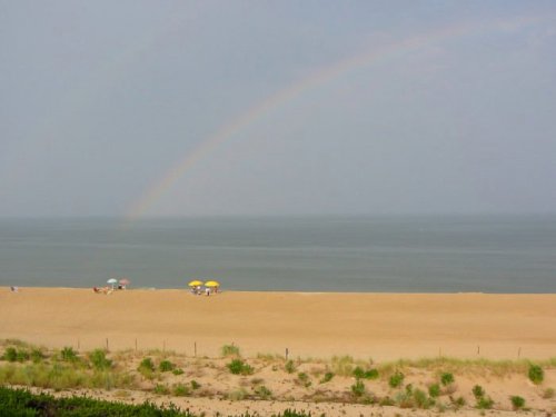 A sandy beach with colorful umbrellas and a faint rainbow over the calm ocean under a cloudy sky.