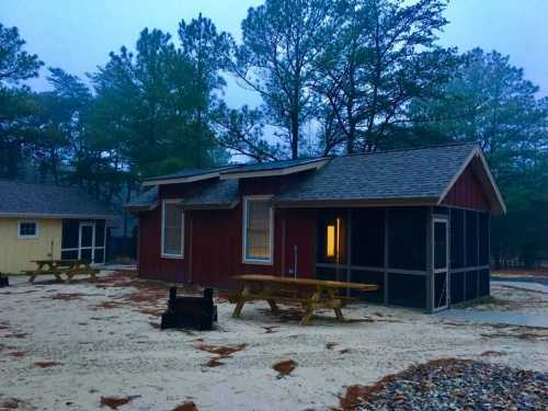 A red cabin with a screened porch surrounded by trees, picnic tables, and a fire pit on a sandy area.