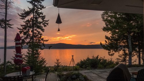 Sunset over a calm lake, framed by trees and a patio, with a camera on a tripod in the foreground.
