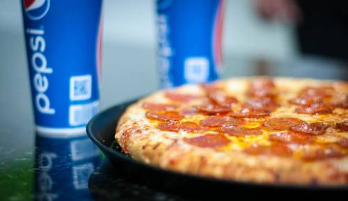 A pepperoni pizza on a black plate with two cups of Pepsi in the background.