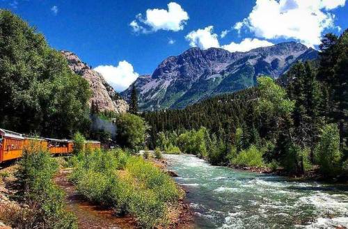 A scenic view of a train alongside a river, surrounded by lush greenery and mountains under a blue sky with clouds.