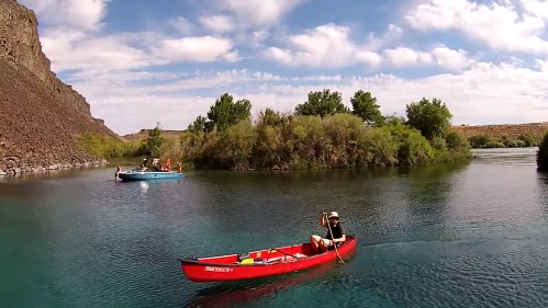 A person paddles a red canoe on a calm river, with a blue boat and lush greenery in the background under a cloudy sky.