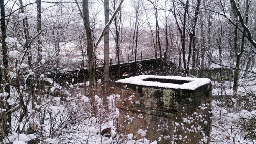 A snowy landscape with a stone structure in the foreground and a wooden bridge in the background, surrounded by bare trees.