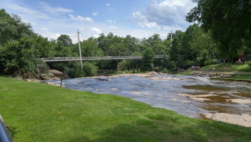 A serene river scene with a footbridge, lush greenery, and a clear blue sky in the background.