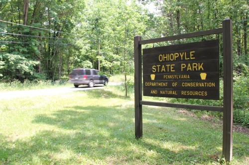 Sign for Ohiopyle State Park in Pennsylvania, with a road and a vehicle passing by in a wooded area.