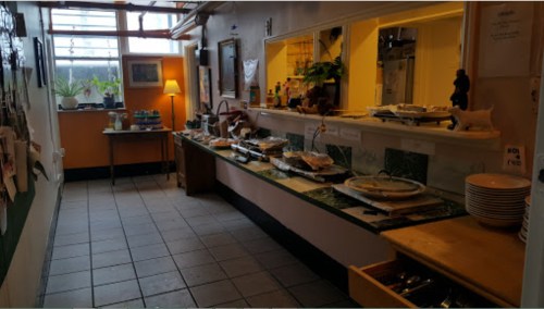 A cozy buffet area with various dishes, a table, and a lamp, set in a well-lit room with plants and artwork.