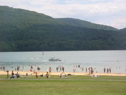 A beach scene with people swimming and relaxing on the sand, surrounded by green hills and a boat on the water.