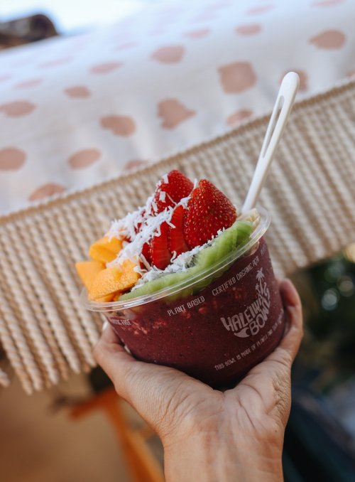 A hand holding a smoothie bowl topped with strawberries, mango, and coconut flakes, with a patterned tablecloth in the background.