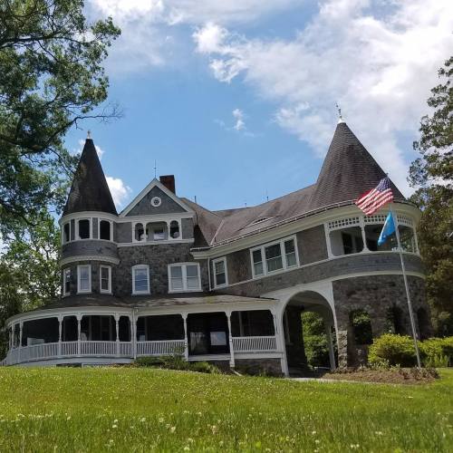 A large, historic stone house with a tower, surrounded by trees and a lawn, featuring American and state flags.