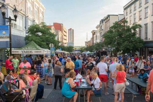 A lively street festival with crowds, food stalls, and people enjoying the event on a sunny day.