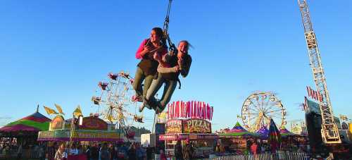 Two women enjoy a zip line ride at a vibrant fair, with colorful booths and a Ferris wheel in the background.