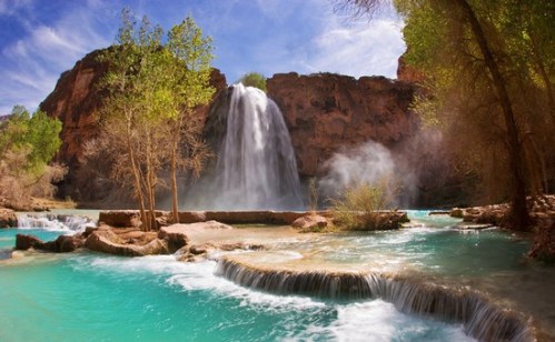 A stunning waterfall cascades into a turquoise pool, surrounded by lush greenery and rocky cliffs under a blue sky.