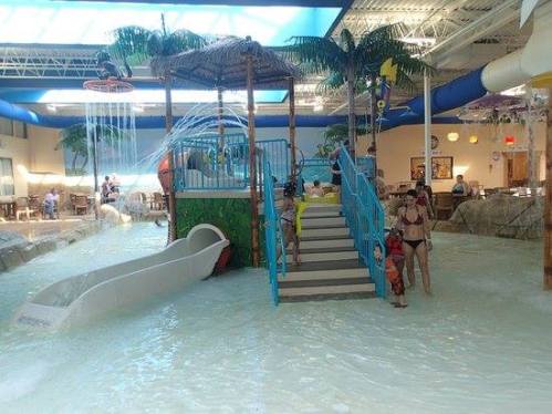 Indoor water park scene with a slide, palm trees, and families enjoying the water. Bright and playful atmosphere.