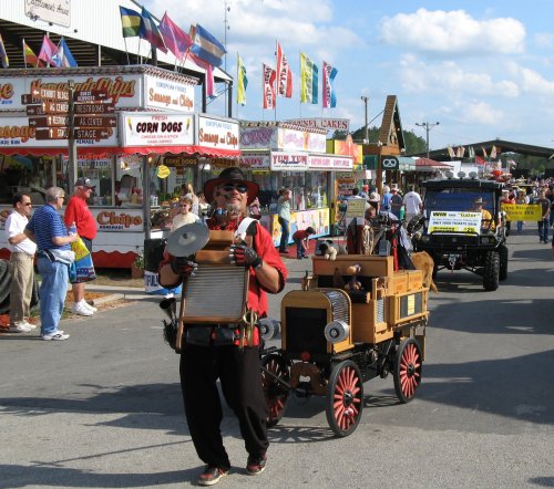 A performer with a washboard and megaphone leads a parade, followed by a small, decorated vehicle at a fair.