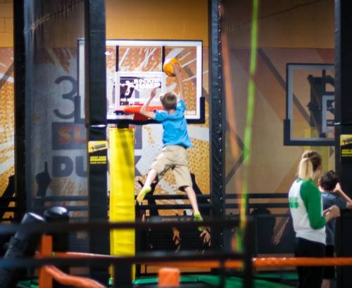 A child jumps to dunk a basketball at a trampoline park, with colorful walls and other people in the background.