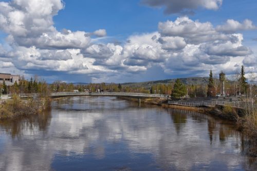 A serene river reflects fluffy clouds and trees under a blue sky, with a bridge in the background.