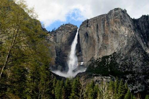 A majestic waterfall cascades down a rocky cliff, surrounded by lush green trees and a cloudy blue sky.