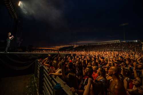 A large crowd at a concert, with a performer on stage and a dramatic sunset in the background.