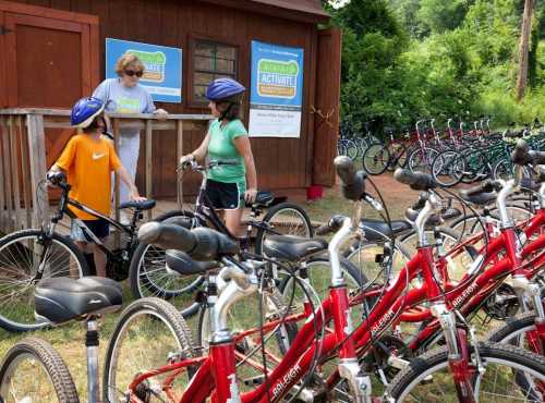 A woman assists two children with bicycles near a shed, surrounded by rows of bikes in a green outdoor setting.