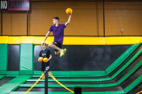 A person jumps on a trampoline while holding a dodgeball, with another person standing nearby in a trampoline park.