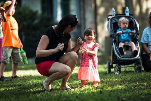 A woman crouches and dances with a smiling toddler in a park, while another child sits in a stroller nearby.