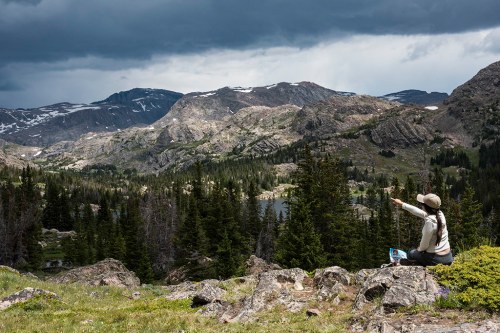 A person sits on rocks in a mountainous landscape, pointing towards distant peaks under a cloudy sky.