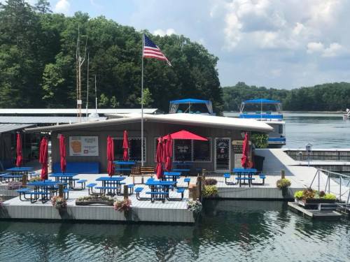 A lakeside restaurant with blue tables and red umbrellas, surrounded by water and trees, featuring an American flag.