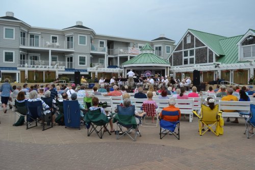 A crowd sits in folding chairs, watching a live band perform in a plaza surrounded by buildings.