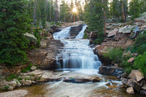 A serene waterfall cascades over rocky steps, surrounded by lush green trees and bathed in warm sunlight.