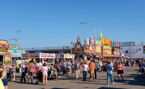 A bustling fairground with food stalls, colorful flags, and crowds enjoying a sunny day.