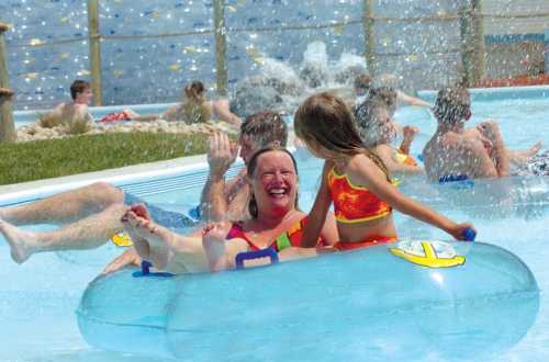 A woman and a girl smile while floating on a blue inner tube in a water park, surrounded by splashing water and other people.