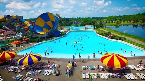 A vibrant water park scene with a large wave pool, colorful umbrellas, and water slides under a blue sky.