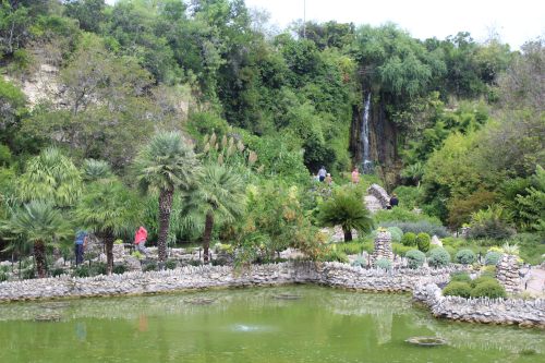 Lush garden with a pond, waterfall, and people walking among greenery and stone pathways.