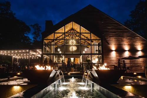 A modern building with large windows, illuminated at night, featuring a water fountain and outdoor seating area.