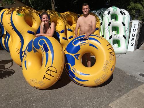 Two people stand with large yellow inner tubes, smiling in front of a backdrop of more tubes and a sign that says "TUBES."