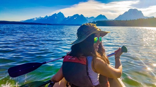 A person in a kayak wearing sunglasses and a hat, paddling on a lake with mountains in the background.