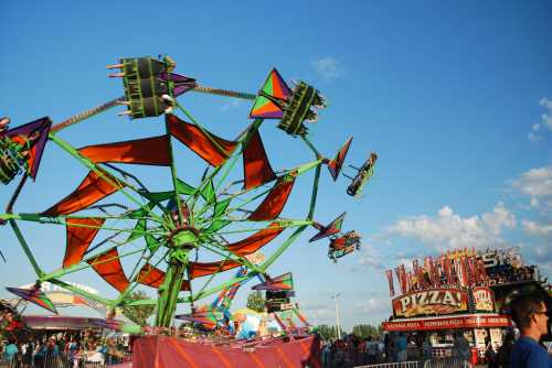 A colorful carnival ride spins against a blue sky, with a pizza stand in the background.