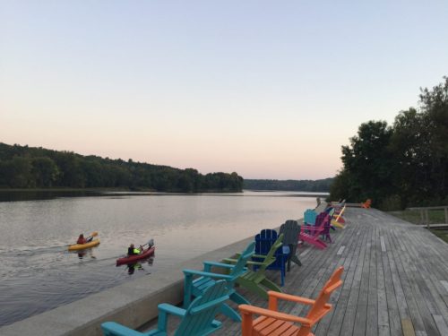 Colorful adirondack chairs line a wooden dock by a calm river, with kayakers paddling in the water at sunset.